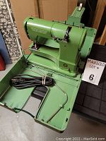 Front view of green Elna sewing machine in an open built-in carrying case, showing power cord, foot pedal socket and metal rod accessory.