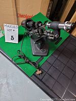 Side view showing the full lensometer on green felt with visible base, eyepiece, dials, and power cord.