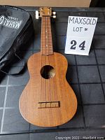 Front view of the Mahalo ukulele with natural wood finish showing the body, neck and visible tuning pegs. The soft case is partially visible.