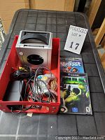 Nintendo GameCube console, cables, controllers, and two game cases all placed inside a red container on a black surface with a MaxSold lot identification card beside it.