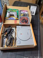 PlayStation system and accessories in open box showing console, cables, new sealed controller, and multiple game discs.