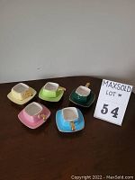 Photo of five espresso cups in different pastel colors each placed on a matching square saucer, arranged in a row displaying the top and side views. All handles are gold colored.