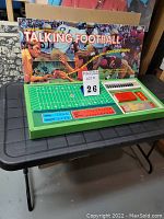 Wide view of the game set on a table with box displayed behind, showing the field game board with blue and red labeled panels, and the box cover art depicting football players in action.