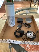 Three Amazon Echo smart speakers in a cardboard box, including a tall white Echo and two Echo Dots (black and white) with power cables.