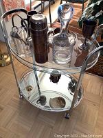 Full view of the round metal frame bar cart with glass top shelf and mirrored bottom shelf, holding glass decanter, metal shaker, glass ice bucket with handle, metal goblet, and other bar items.