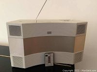Front angled view of Bose Acoustic Wave Radio system showing speaker grilles, beige and tan color scheme, and attached remote control on front.