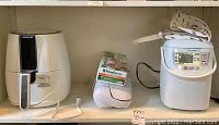 Shelf display photo showing three white kitchen appliances: air fryer, vacuum sealer, and bread maker with cloth cover.