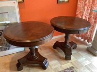 Pair of matching round wood side tables placed side-by-side against an orange wall, showing the tops and bases.