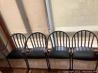 Front view of four metal armchairs showing curved armrests, vertical slat backrests, and padded black seats.