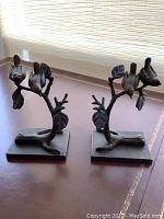 Pair of bronze bird figurines shown on surface near window. Each piece depicts birds sitting on detailed tree branches with leaves, mounted on rectangular bases.
