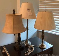 All three table lamps on a dark wooden surface near window with blinds: dark wood base with beige bell-shaped shade, tall silver base with white tapered shade, brass base with cream pleated shade.