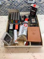 Box of multiple tobacco related items including various lighters, Ronson butane fuel can, Zippo lighter fluid can, and accessories