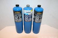 Photo of three blue Bernzomatic propane cylinders, two with black caps and one without, all labeled with safety warnings and brand information.