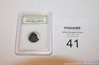 Front view of the slabbed ancient Roman coin with label showing era and registry number, alongside auction identifier card.