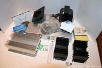 Mixed lot of electronic items arranged: large and small heat sinks, small cooling fan, analog meters, plastic hardware parts, metal enclosure box.