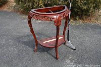 Full view of the carved wooden accent table showing half-moon top frame and curved legs outdoors with hockey stick for scale.