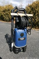 Front view of blue and black Simoniz pressure washer with hose on reel and nozzle attached to handle.