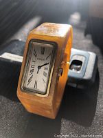 Photo showing front and slight angle of vintage Agon watch with yellow bakelite bangle on a dark surface with a mobile phone in background.