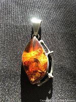 Amber and sterling silver pendant photographed on black fabric, showing front and side profile with visible inclusions inside the amber cabochon.