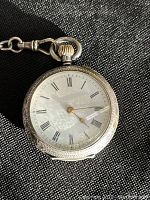 Front of pocket watch showing white dial with black Roman numerals and gold tone hands in engraved sterling case
