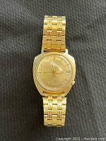 Full view of the vintage Bulova Accutron watch lying flat on dark fabric, showing the gold tone bracelet and watch face with date display at 3 o'clock.