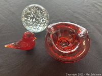 Group photo of three items: clear bubble paperweight, red glass bird figurine, and red glass bowl, displayed on black fabric.