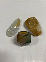 Three items visible on white fabric surface: two carved jade pieces with brownish-orange translucent hues and one multi-colored agate ring with angular facets.