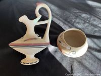 Two vintage pottery pieces on black fabric background: a handled pitcher and a small rounded bowl, both decorated with feather and floral designs.