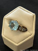 Ring on white leatherette display at slight angle showing front view of octagonal blue topaz and right shoulder accents