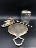 View showing entire lot with the mirror facedown, two glass jars with lids, the button hook, and lint brush arranged on a black background.