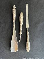 Three sterling silver items displayed against a black background: a shoe horn, a file, and a scraper, showing the varying sizes and detailed handle designs.