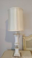 Photo of one cream and gold table lamp with tall cylindrical white shade on cream nightstand beside cane back chair