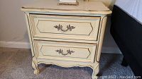 Photo of one yellow and gold wooden end table showing two drawers with ornate handles, painted with a distressed finish and carved decorative legs.