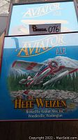 Photo showing two framed Aviator Ale signs with seaplane artwork, seller inventory tag 076 attached.