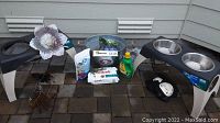Lot arranged on patio, showing two elevated pet feeders with stainless steel bowls, bird feeder, metal tub, plant fertilizers, bird food, liquid fertilizer, and wind chime.