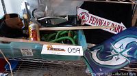 Photo shows a pair of black folding stadium seats, brown leather tool belt, various brushes, green flexible garden hose inside a plastic bin, can of WD-40 lubricant, plastic funnel, tape rolls, Melnor sprinkler, and Boston Fenway Park metal sign with logo behind them. A Seattle Mariners flag is visible draped underneath the sign.