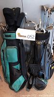 Two golf bags with clubs standing side by side; left bag is teal and black, right bag is black with gray and blue accents.