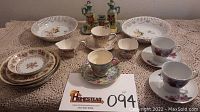 All items displayed on crocheted tablecloth including porcelain cups, saucers, plates, bowls, and cruet set.