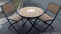 Photo showing two folding chairs and round table arranged outdoors on pavement.