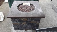 Square propane fire pit with faux stone base and wooden-look top, round fire bowl filled with lava rocks.