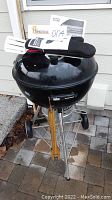Weber kettle-style charcoal grill with black lid and wooden handle on vent, shown outside on patio