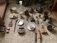 Full layout of assorted antique household items on carpet including teapots, glass bottles, utensils, small tools, and decorative ceramics