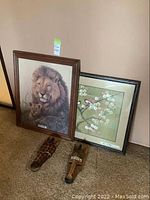 Two framed prints on the floor leaning against wall: lion and cub art, floral and bird art, with two animal masks beneath.