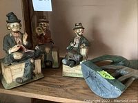 Three distinct porcelain clown music boxes in seated poses with detailed clothing and accessories on wooden bases.