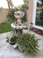 Full view of two-tier beige cast stone fountain with floral accents, surrounded by potted succulents and decorative plants on concrete patio outside home.