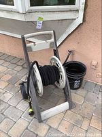 Front angle view of the hose holder cart with wound garden hose and black bucket next to it on paved ground against a beige house wall.