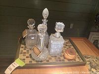 Four clear glass liquor decanters with stoppers on a rectangular tile and wood tray with three round natural wood coasters next to them.