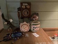 Photo showing an old style rotary phone mounted on a wall, eagle sculpture, small clock, dream catcher, ceramic sake barrel, and a decorative feather headdress on a wooden surface.