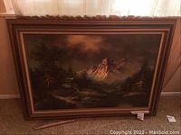 Framed landscape canvas painting showing mountain peak and pine trees in moody lighting, photographed leaning against a wall.