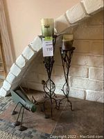 Photo of three metal candle holders of different heights with green pillar candles, plus metal cricket figurine in front of them next to a brick fireplace.